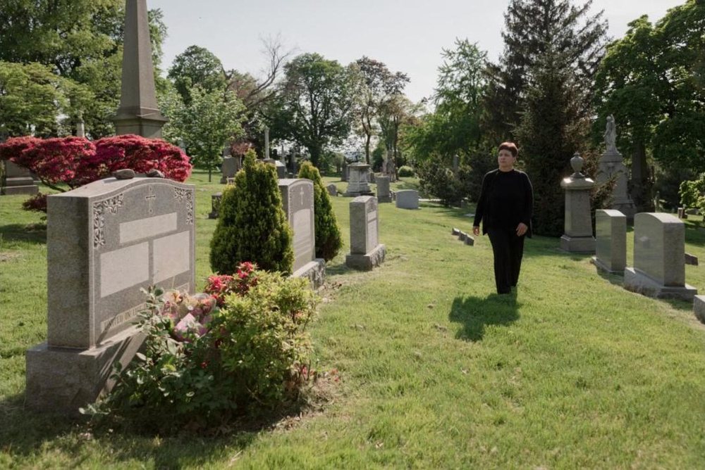 A woman walking across a sunny, green cemetery to the next grave